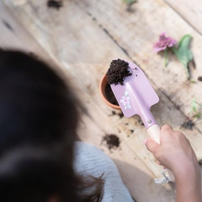 Pessoa segurando pá de jardinagem lilás com terra sobre vaso de barro pequeno