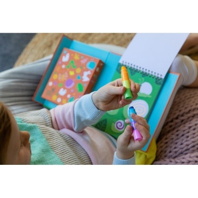 Criança segurando canetas coloridas e a desenhar em caderno aberto