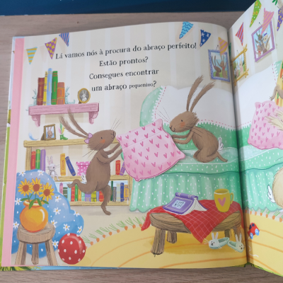 Ilustração colorida de livro infantil com duas lebres castanhas a brincar numa cama verde, texto em português e cenário decorado com livros, flores, mesa e almofadas.