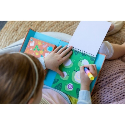 Criança a desenhar num caderno espiral com caixa azul e livrinho laranja ao lado