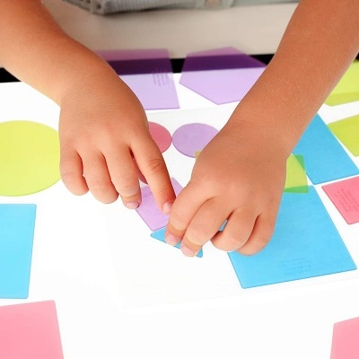 Criança a montar formas geométricas coloridas numa mesa de luz branca.