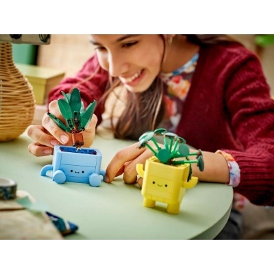 Brinquedos de montar vasos sorridentes azuis e amarelos com plantas verdes numa mesa verde clara com menina sorridente ao fundo.