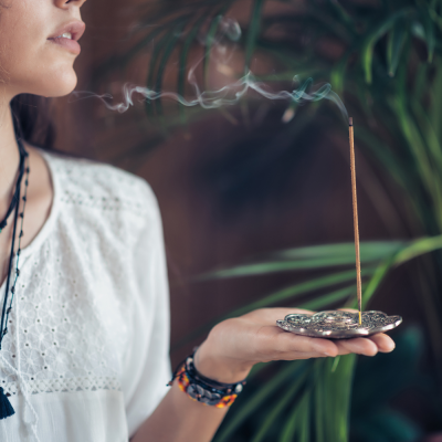 Incenso aceso num suporte metálico em forma de flor na mão de uma pessoa com blusa branca e plantas ao fundo
