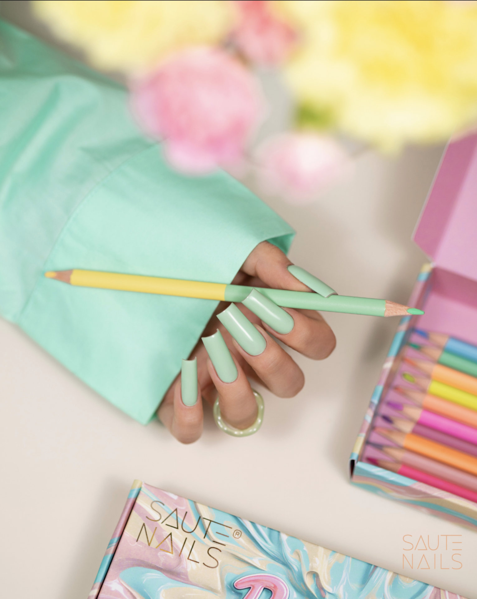 Verniz Gel Saute Nails S295 - Minty Chill (Verde-Menta Pastel) Mão com unhas verdes segurando lápis de cor ao lado de caixa com lápis coloridos