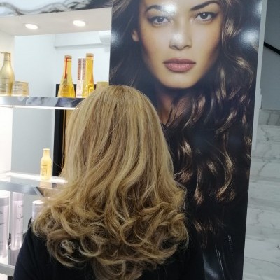 Pessoa de costas com cabelo loiro ondulado à frente de cartaz de mulher com cabelo castanho ondulado e prateleira de produtos L'ORÉAL