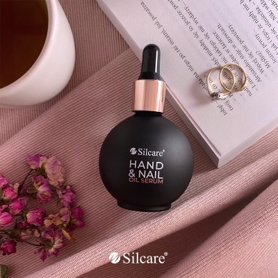 frasco preto redondo com conta-gotas dourada de óleo para mãos e unhas Silcare sobre tecido rosa ao lado de livro, anéis, flores secas e chávena de chá