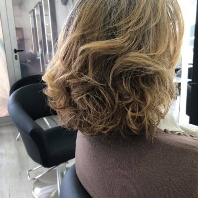 Cabelo loiro ondulado e volumoso numa pessoa sentada numa cadeira preta de salão de cabeleireiro