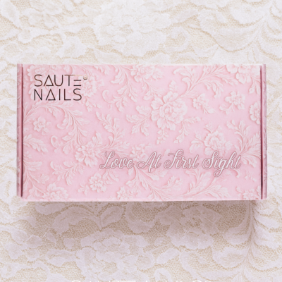 Embalagem rosa com padrão floral e texto SAUTE NAILS e Love At First Sight sobre tecido bege