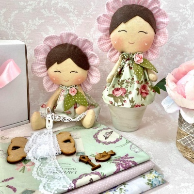 Duas bonecas de tecido com vestidos florais, entre tecidos dobrados, caixa branca com fita rosa e flor rosa.