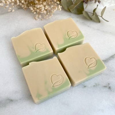 Barras de sabão bege e verde com símbolo de coração em superfície branca e flores secas ao fundo