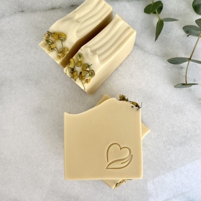 Duas barras de sabão creme com flores secas e símbolo em relevo, sobre mármore branco e folhas verdes