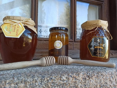 Três frascos de mel com diferentes etiquetas e tampas, sobre balcão de pedra com colheres de mel