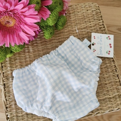 Cuecas para bebé xadrez azul e branco sobre tapete de vime com flores cor-de-rosa ao lado