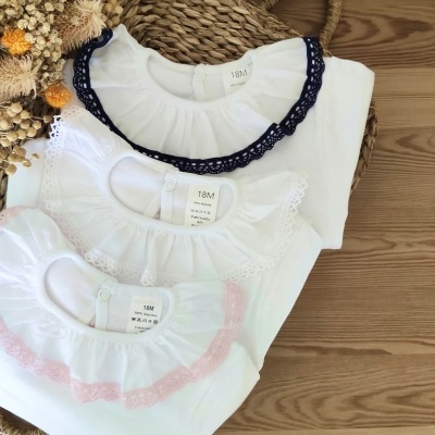 Três bodies de bebé brancos com golas de renda coloridas em cima de superfície de madeira perto de cesto e flores secas