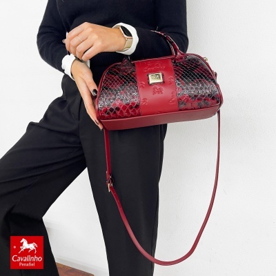 Mala vermelha em pele com padrão tipo pele de réptil e detalhes lisos, com logótipo metálico e alça longa, segurada por mulher vestida de preto