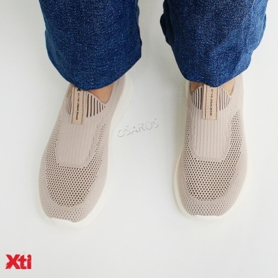 Sapatilhas bege em tecido de malha com sola branca e etiqueta na língua, usadas com calças de ganga azuis.