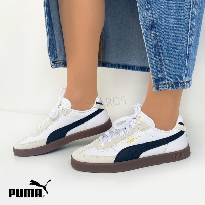 Sapatilhas Puma brancas com detalhes castanhos e azul escuro, usadas com calças de ganga azul.