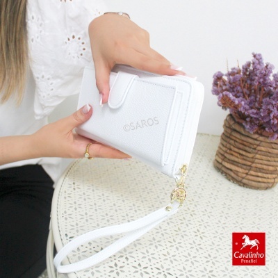 Carteira branca em couro com textura e fecho de correr, com alça de pulso, sobre mesa branca com vaso de flores roxas