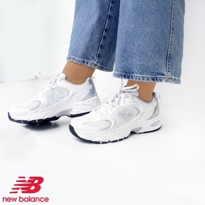 Ténis brancos New Balance com sola preta e branca usados com calças de ganga azuis.