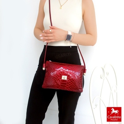 Mala vermelha com padrão pele de cobra e fecho dourado, usada por mulher com roupas brancas e pretas, cadeira branca ao lado