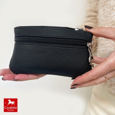 Carteira preta pequena com fecho de correr e puxador metálico, segurada por duas mãos femininas com unhas castanho escuro.