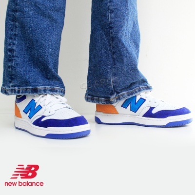 Sapatilhas New Balance brancas com detalhes azuis e laranja, usadas com jeans azuis, em fundo branco