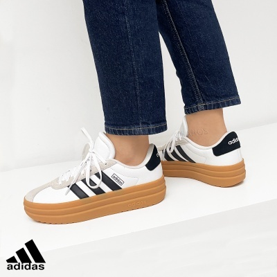 Ténis Adidas branco com riscas pretas e sola castanha, usados com jeans.