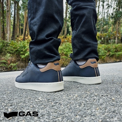 Ténis em pele azul escuro com detalhes castanhos e sola branca, numa estrada exterior com vegetação ao fundo.