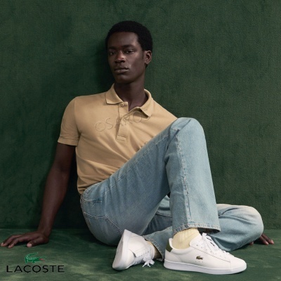 Homem sentado com polo bege, jeans azuis claros e sapatilhas brancas Lacoste em fundo verde.