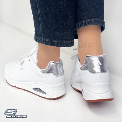 Sapatilhas brancas Skechers com detalhe prateado no calcanhar, sola branca e castanha, usadas com calças de ganga azuis.