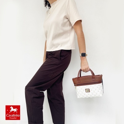 Mulher com camisola bege segura mala branca com detalhes castanho em fundo branco