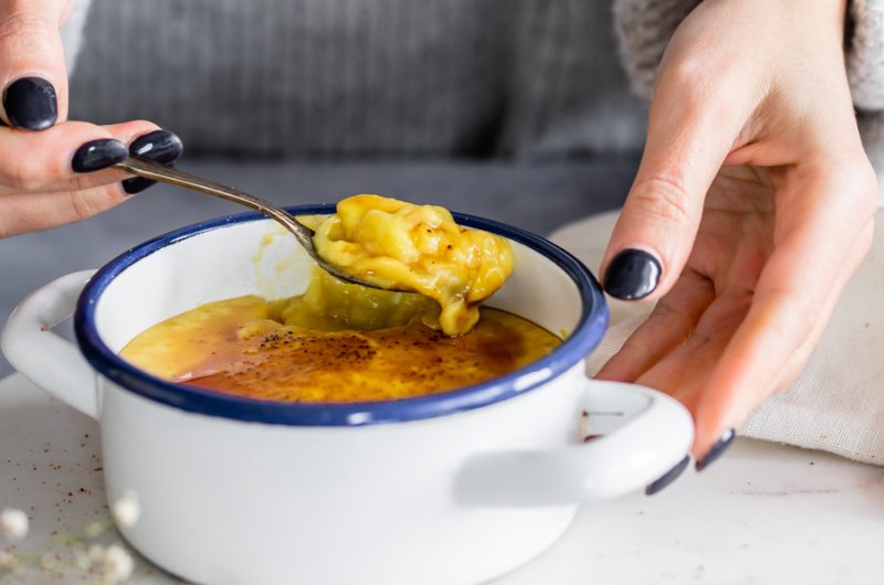 Creme amarelo queimado numa taça branca com bordo azul escuro, com colher e mãos de unhas pretas.