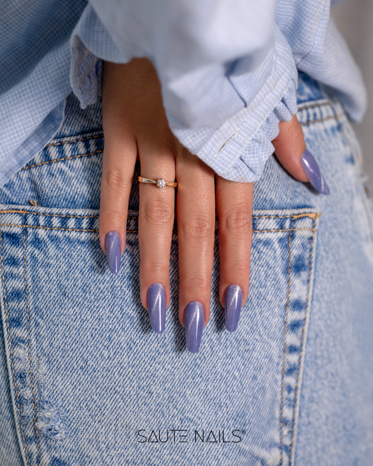 Verniz Gel Cateye Effect E14 (Azul com um Brilho Prateado) Mão feminina com unhas lilás cintilantes e anel, apoiada em calças de ganga azul.
