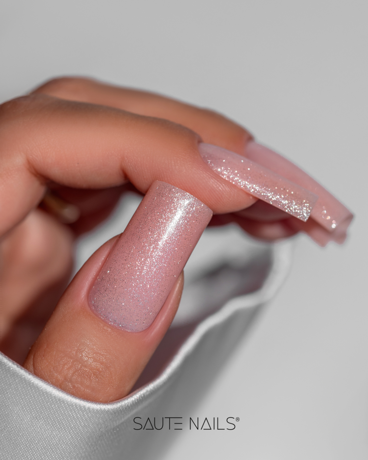 Verniz Gel S316 - Soul Touch (Bege com Partículas e Reflexos Multicoloridos) Mãos com unhas rosa cintilante segurando tecido branco com logótipo Saute Nails