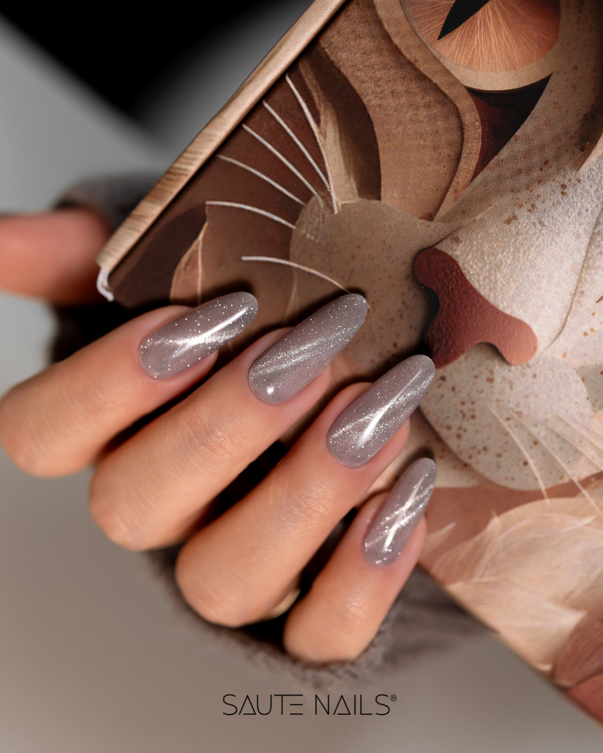 Verniz Gel Cateye Effect E16 (Cinzento com Brilho Prata) Unhas artificiais cinzentas com glitter prateado em formato almendrado segurando caixa com ilustração artística