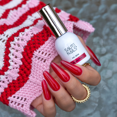 Unhas vermelhas brilhantes com verniz Saute Nails segurando frasco branco sobre malha vermelha e rosa.