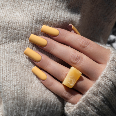 Mão com unhas amarelas muito compridas e anel retangular amarelo sobre malha bege tricotada.