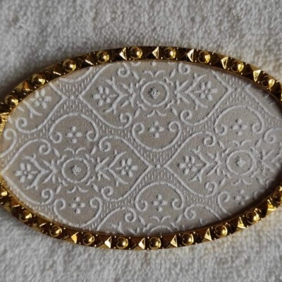 Objeto oval branco com padrão decorativo e borda dourada com detalhes rebaixados