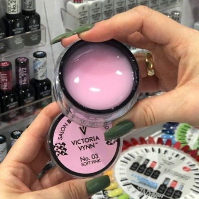 Mãos segurando gel para unhas rosa da Victoria Vynn com expositor de esmaltes e amostras ao fundo