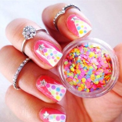 Mão com unhas decoradas em rosa, branco e pontos coloridos, anéis prateados e frasco de confetes coloridos
