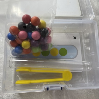 Conjunto em caixa transparente com bolas coloridas, pinça amarela e cartão com círculos coloridos