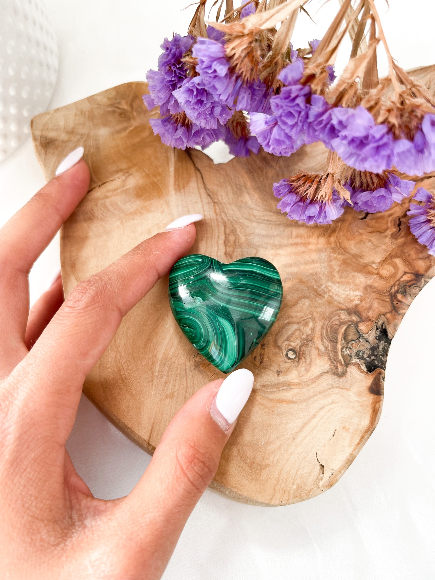 Coração de pedra verde com padrões ondulados numa tábua de madeira com flores secas roxas e mão feminina com unhas brancas