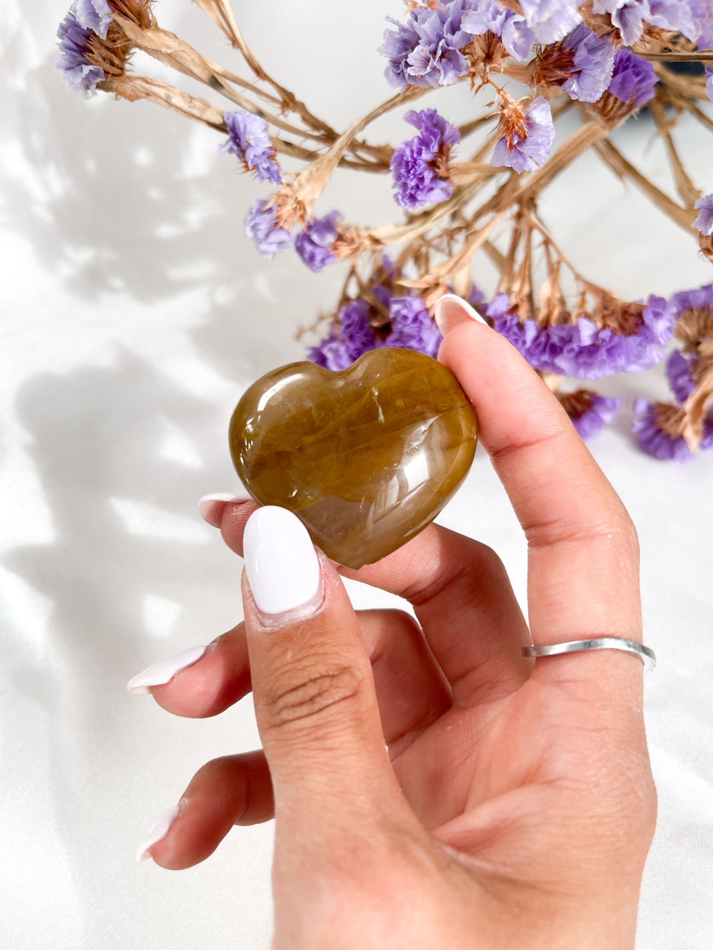 Mão segurando pedra em forma de coração castanha com flores secas lilases ao fundo