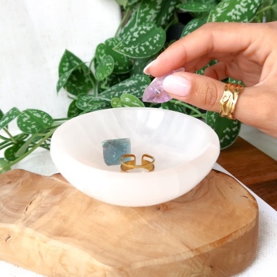 Anel dourado ondinado e pedras coloridas numa mão sobre tigela branca e base de madeira com folhas verdes ao fundo.