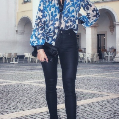 Mulher com camisa azul e branca de padrão abstrato, calças justas pretas e botas, segurando bolsa preta num espaço urbano