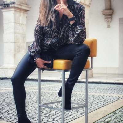 Mulher com blusa animal print sentada num banco amarelo alto em pavimento de pedra com arcos ao fundo