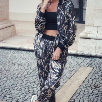 Pessoa com conjunto estampado em tons castanho e preto, top preto e sapatilhas brancas em calçada de pedra.