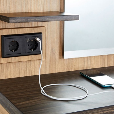 Tomada elétrica dupla embutida numa parede de madeira com cabo branco ligado a telemóvel numa mesa de madeira escura