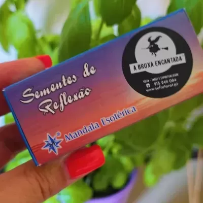 Caixa colorida com texto 'Sementes de Reflexão' e selo 'A BRUXA ENCANTADA', mão com unhas vermelhas a segurá-la, planta verde no fundo