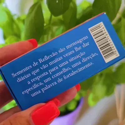 Caixa azul com texto em português segurada por mão com unhas vermelhas em fundo de planta verde e decoração
